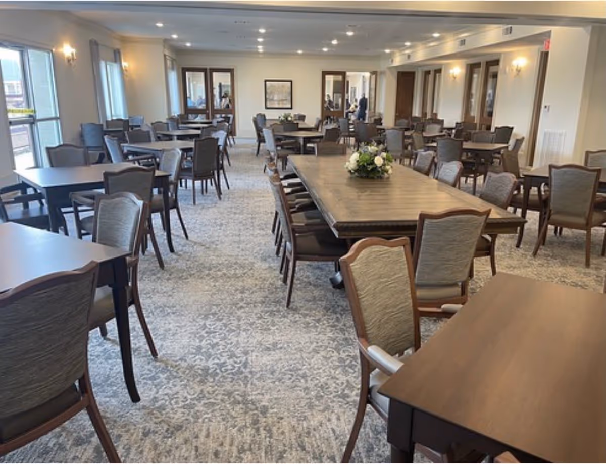 A spacious dining room with multiple wooden tables and cushioned chairs arranged neatly. The room has large windows on the left side allowing natural light to fill the space. There are floral centerpieces on some tables and wall sconces providing additional lighting. The carpet has a subtle patterned design, and the walls are painted in a light color.