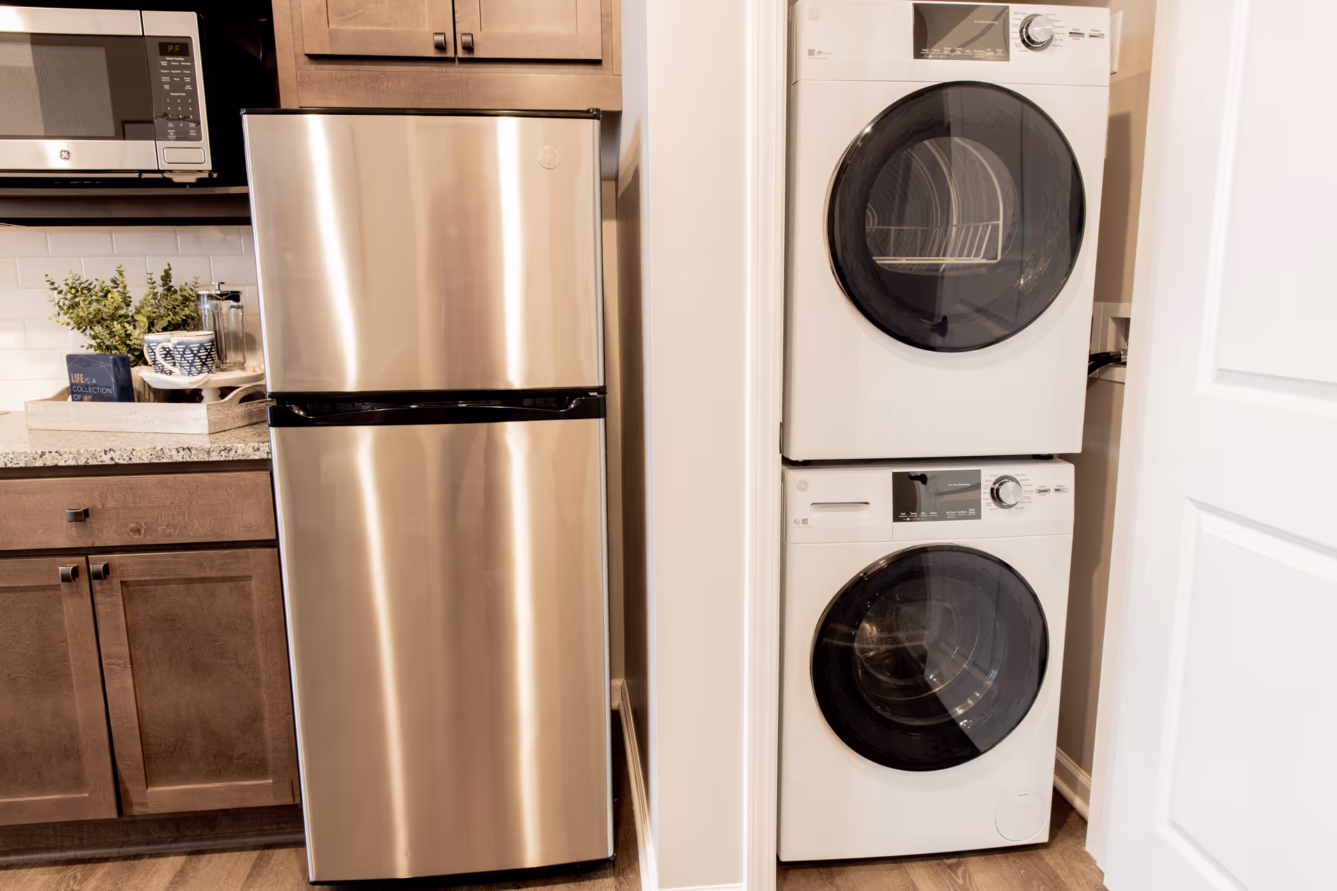 Stainless steel refrigerator next to a closet with a stacked washer and dryer beside kitchen cabinets and countertop.