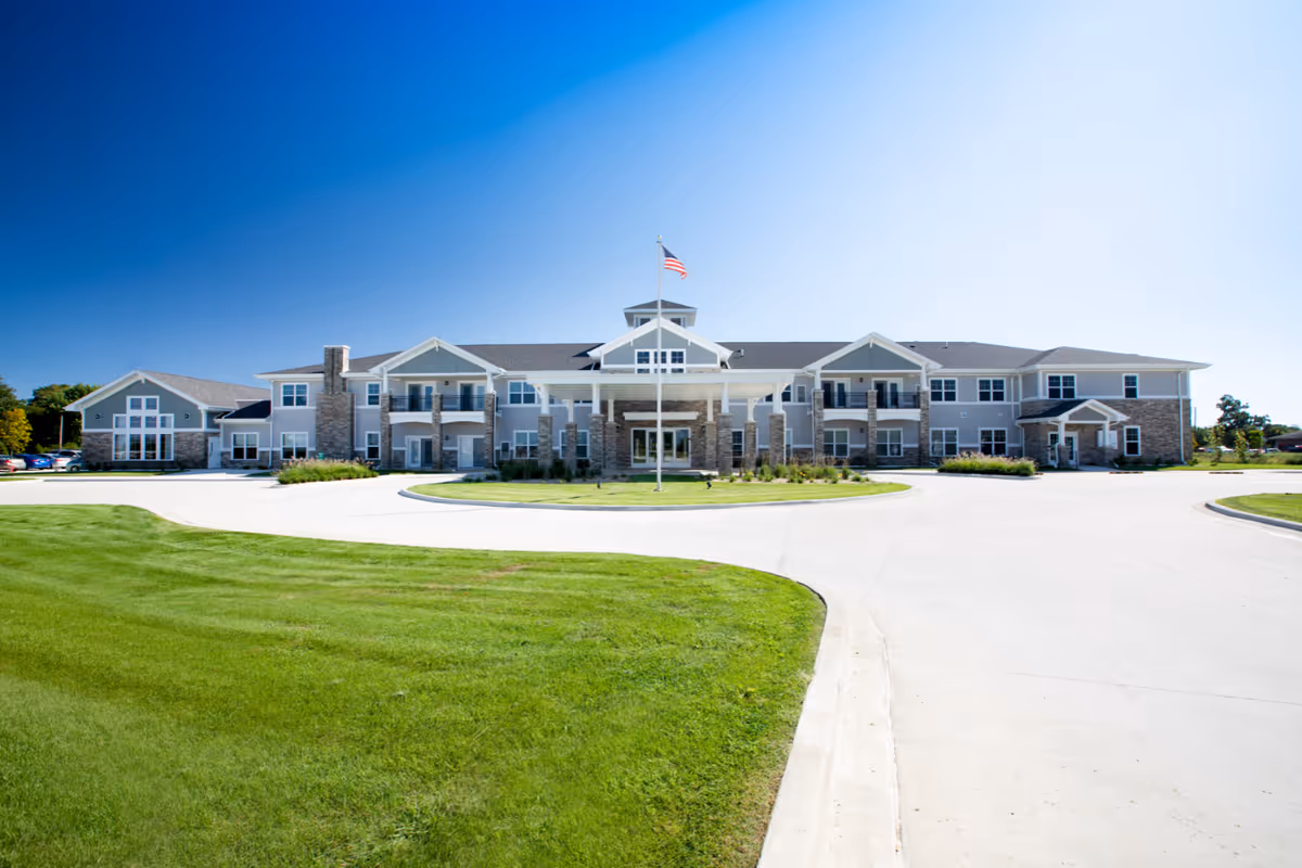 Front exterior view of Cedarhurst Senior Living of Arnold building with a large driveway, green lawn, and an American flag on a flagpole in front under a clear blue sky.