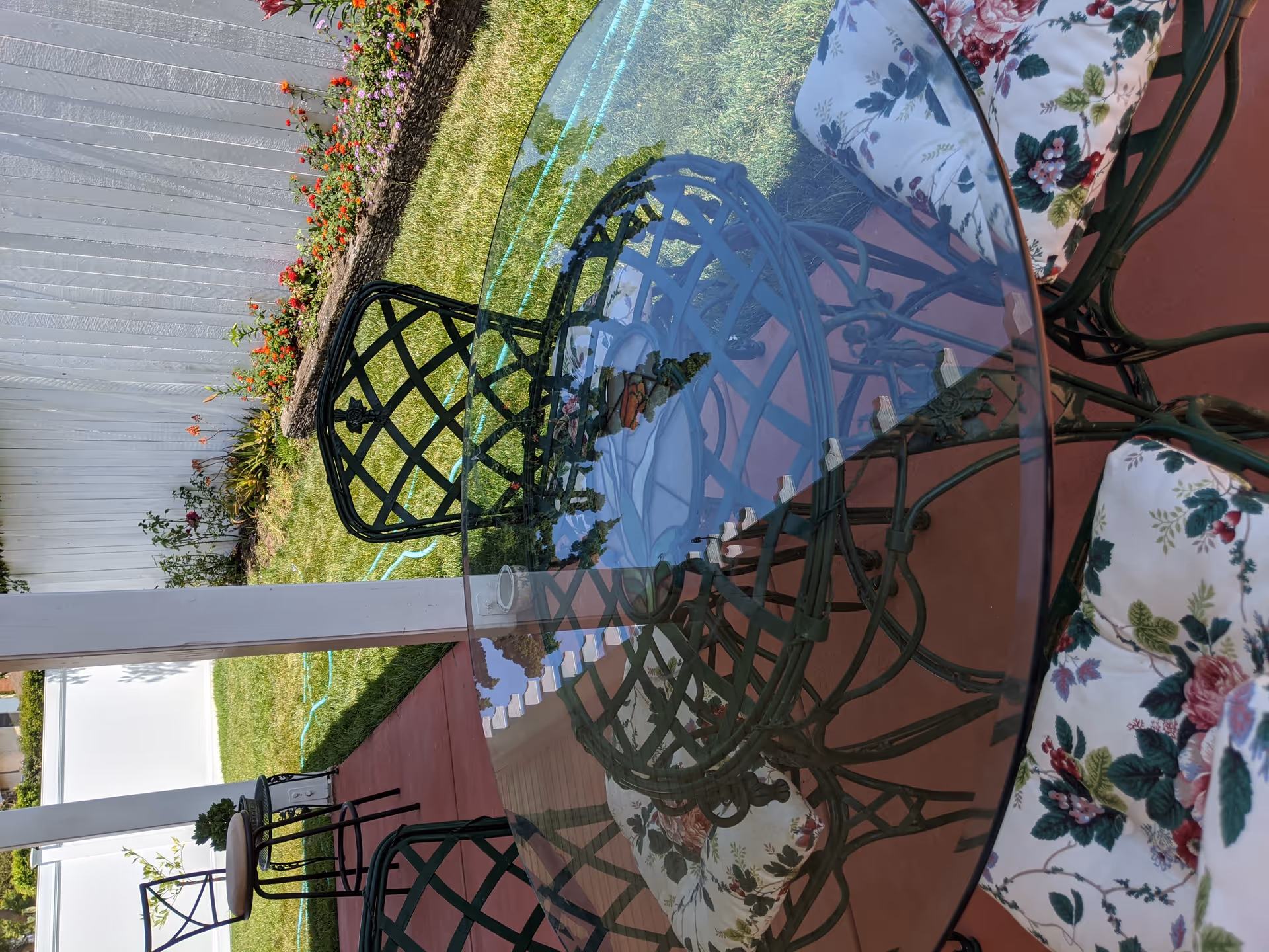 Outdoor patio area with a round glass-top metal table and four metal chairs with floral cushions. The patio has a red concrete floor and is adjacent to a grassy lawn with a white fence and a flower bed along the fence.