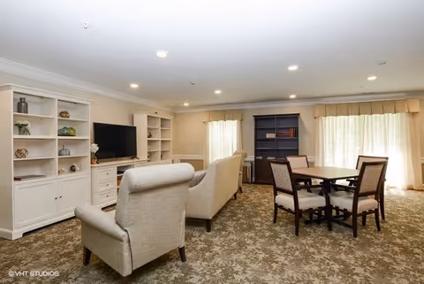 A spacious living room with beige armchairs and a sofa arranged around a large flat-screen TV on a white built-in shelving unit. There is a dark wood dining table with four chairs near large windows covered with light curtains. The room has recessed ceiling lights and a patterned carpet floor.