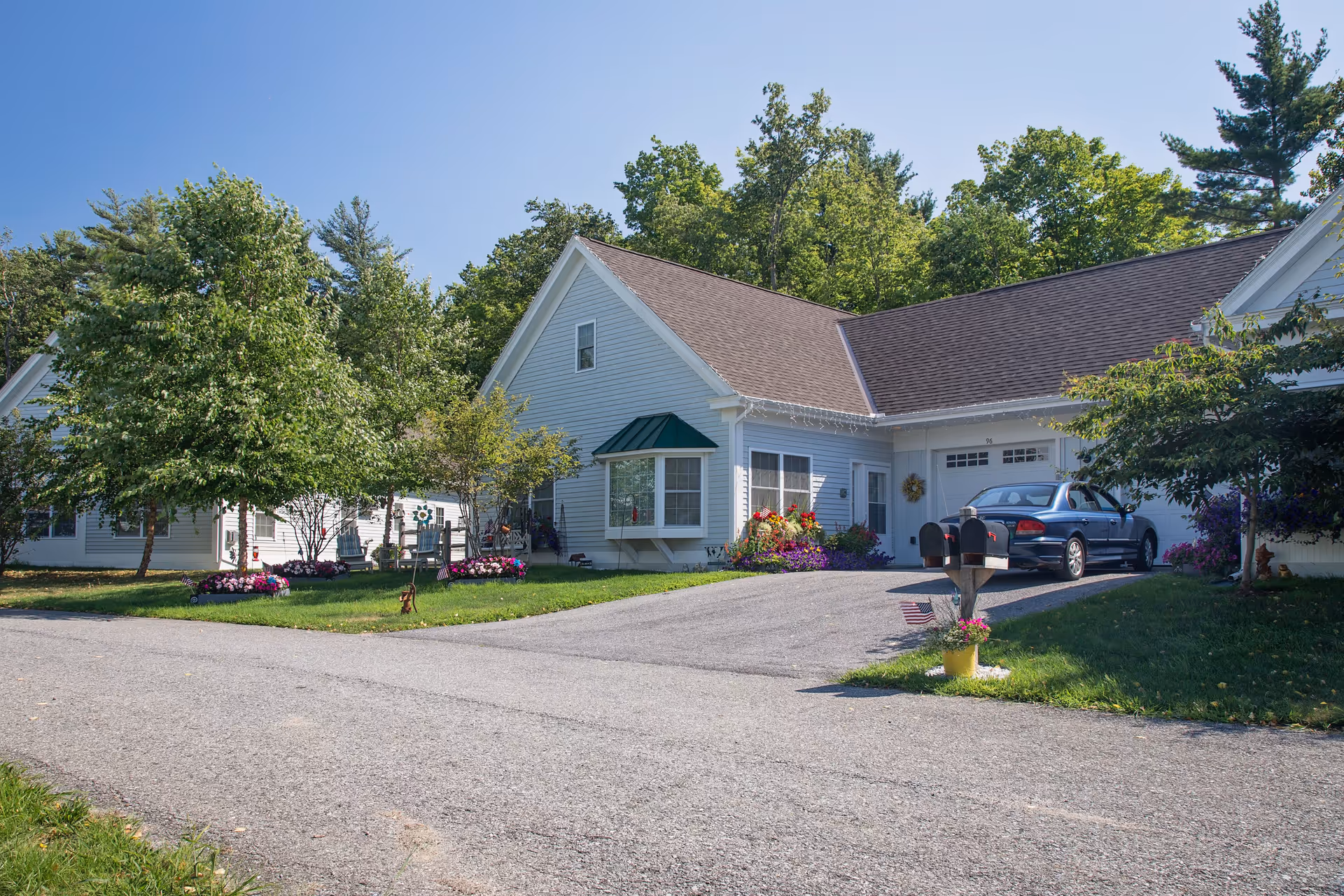 Exterior view of a residential building at The Residence at Otter Creek with a driveway, a parked blue car, green trees, and colorful flower beds under a clear blue sky.