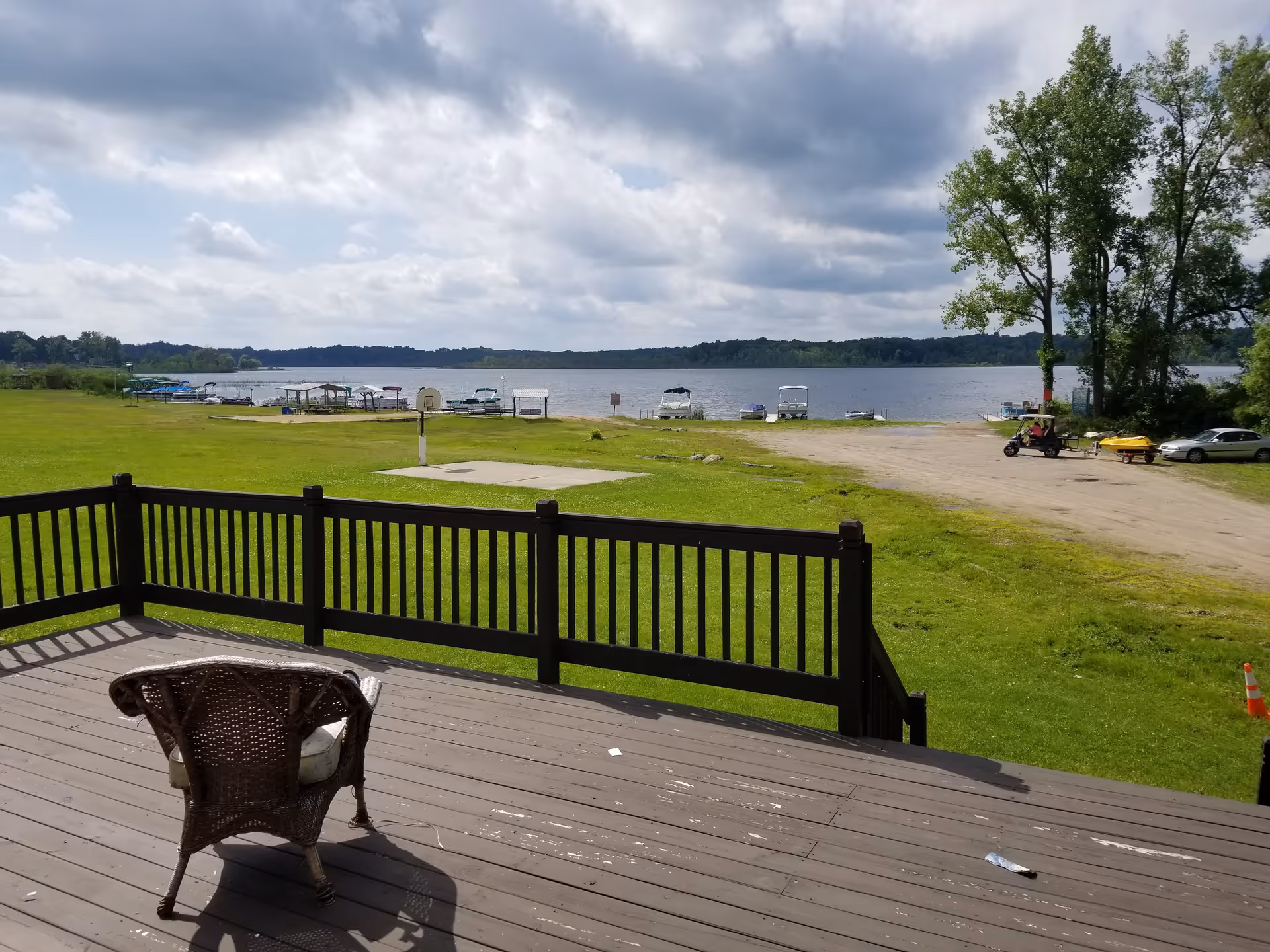 View from a wooden deck with a single wicker chair facing a grassy area leading to a lake with several boats docked along the shore under a partly cloudy sky.
