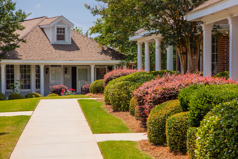 A paved walkway leading to a single-story residential building with a beige roof and white exterior. The building has black shutters and a door labeled 205. Well-maintained bushes and shrubs with green and reddish foliage line the walkway on both sides, and there are trees providing shade.