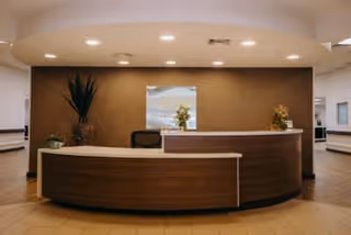 Reception area with a curved wooden desk, a chair behind it, decorative plants on the desk and floor, and a framed picture on the wall with recessed ceiling lights above.