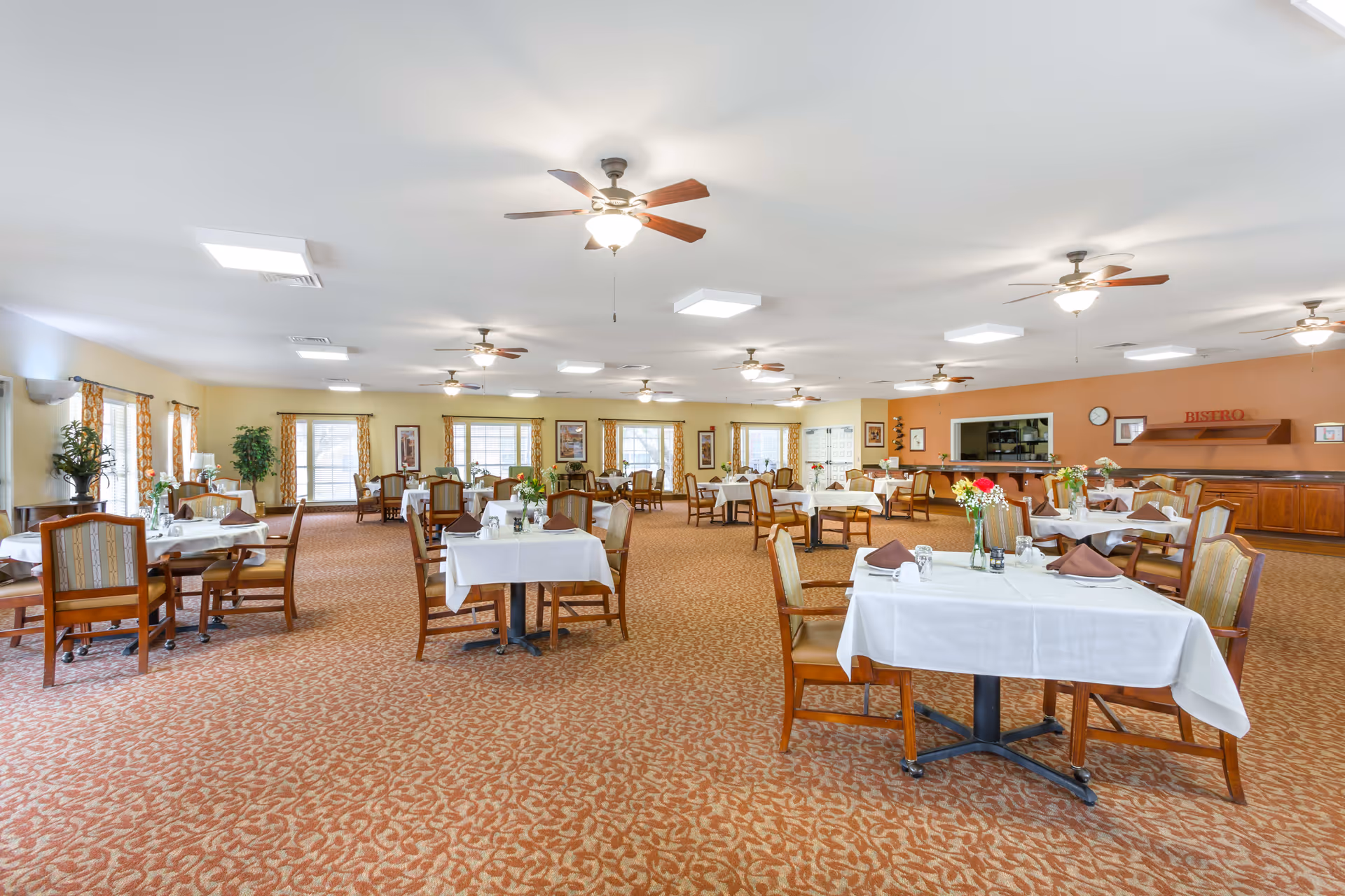 A spacious dining room with multiple tables covered in white tablecloths, each set with napkins, glasses, and flower vases. The room has carpeted floors, ceiling fans with lights, large windows with patterned curtains, and a serving area labeled 'Bistro' in the background.