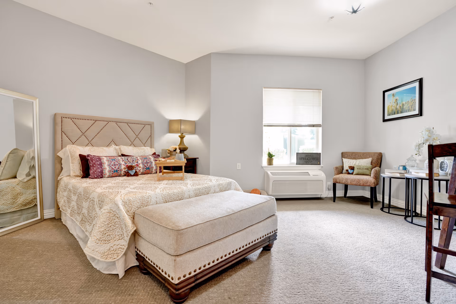 A bright and spacious bedroom with a large bed featuring a beige quilt and decorative pillows. There is a cushioned bench at the foot of the bed, a floor mirror leaning against the wall, a bedside table with a lamp, and a window with blinds letting in natural light. A comfortable armchair with a pillow and a small table with decorative items are placed near the window. The walls are painted light gray and the carpet is beige.