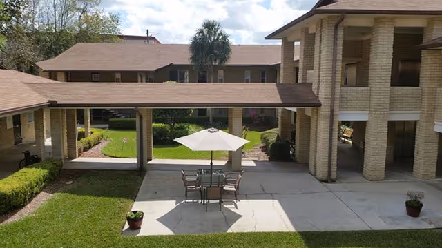 Courtyard of an assisted living facility with a patio table and umbrella, covered walkways, and a two-story building surrounding a lawn.
