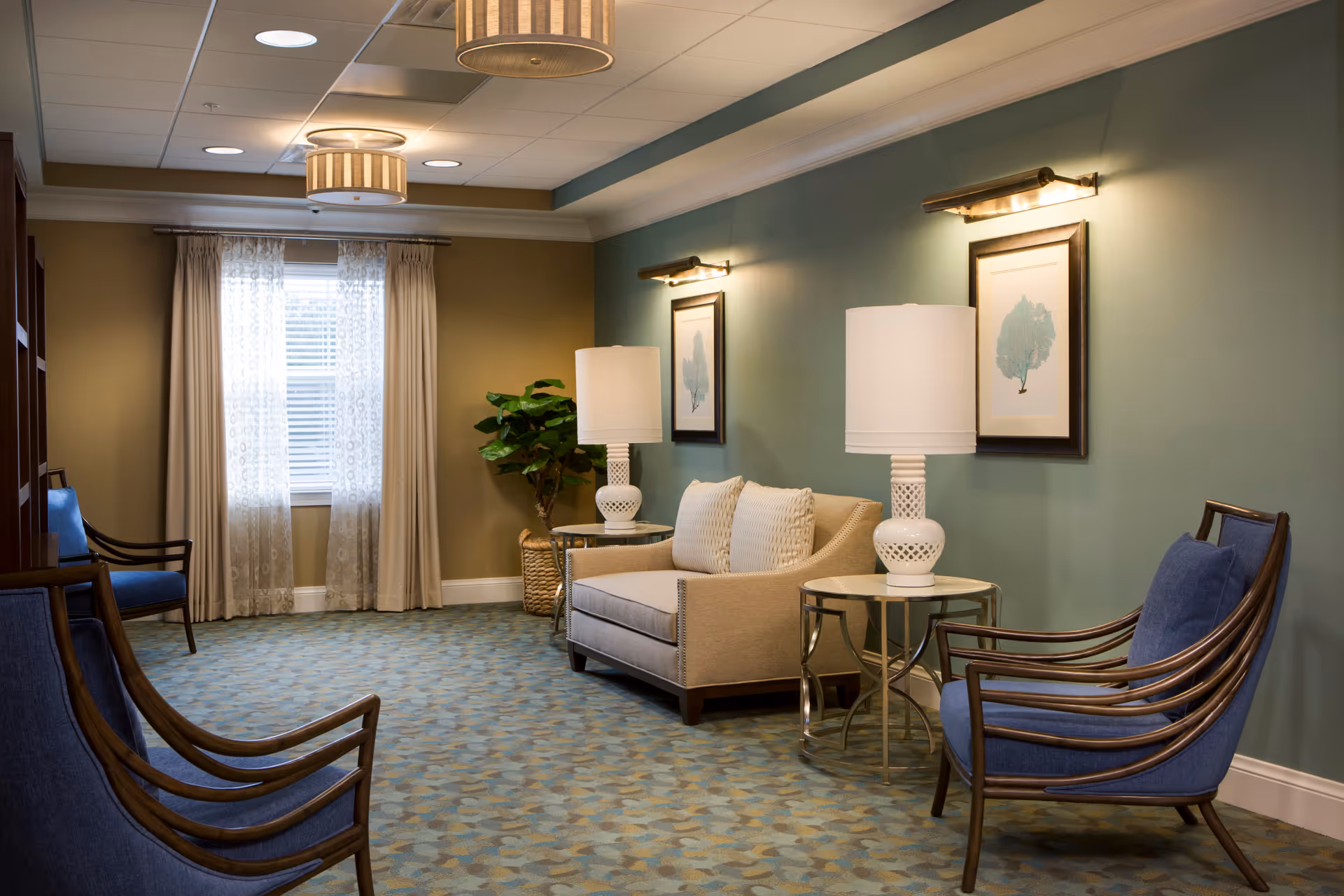 A cozy sitting area in a senior living facility with a beige loveseat, two blue cushioned armchairs, two white table lamps on round side tables, framed artwork on a green wall, a window with sheer curtains, and a potted plant in the corner.