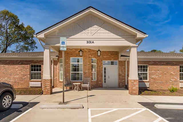 Front exterior view of a single-story brick building with a covered entrance supported by white columns. There are two chairs and a small table under the entrance roof. A handicap parking sign is visible near the entrance, and a car is partially visible on the left side. The sky is clear and blue.