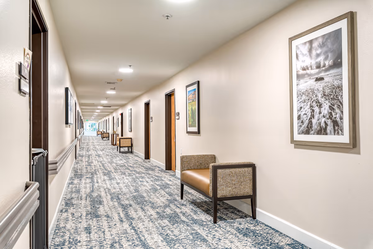 A long, well-lit hallway in a senior living facility with patterned carpet, beige walls, and several framed landscape pictures hanging on the walls. There are cushioned benches placed along the hallway and multiple doorways leading to rooms.