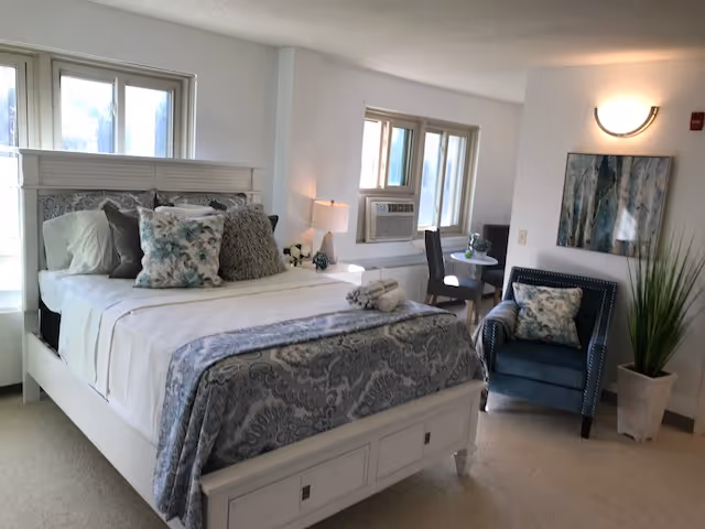 Well-lit furnished bedroom with a white bed dressed in patterned bedding, an upholstered armchair, a small table, and multiple windows.