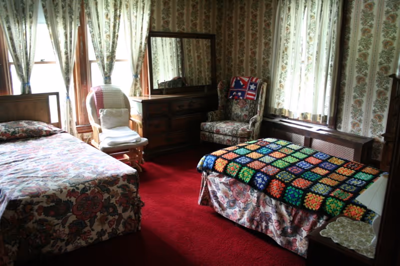 A cozy bedroom with two beds covered in floral bedspreads, one with a colorful crocheted blanket. The room has red carpet, floral wallpaper, a wooden dresser with a mirror, a cushioned armchair with a patriotic quilt draped over it, and a wooden rocking chair with a white cushion. Light filters in through two windows with floral curtains.