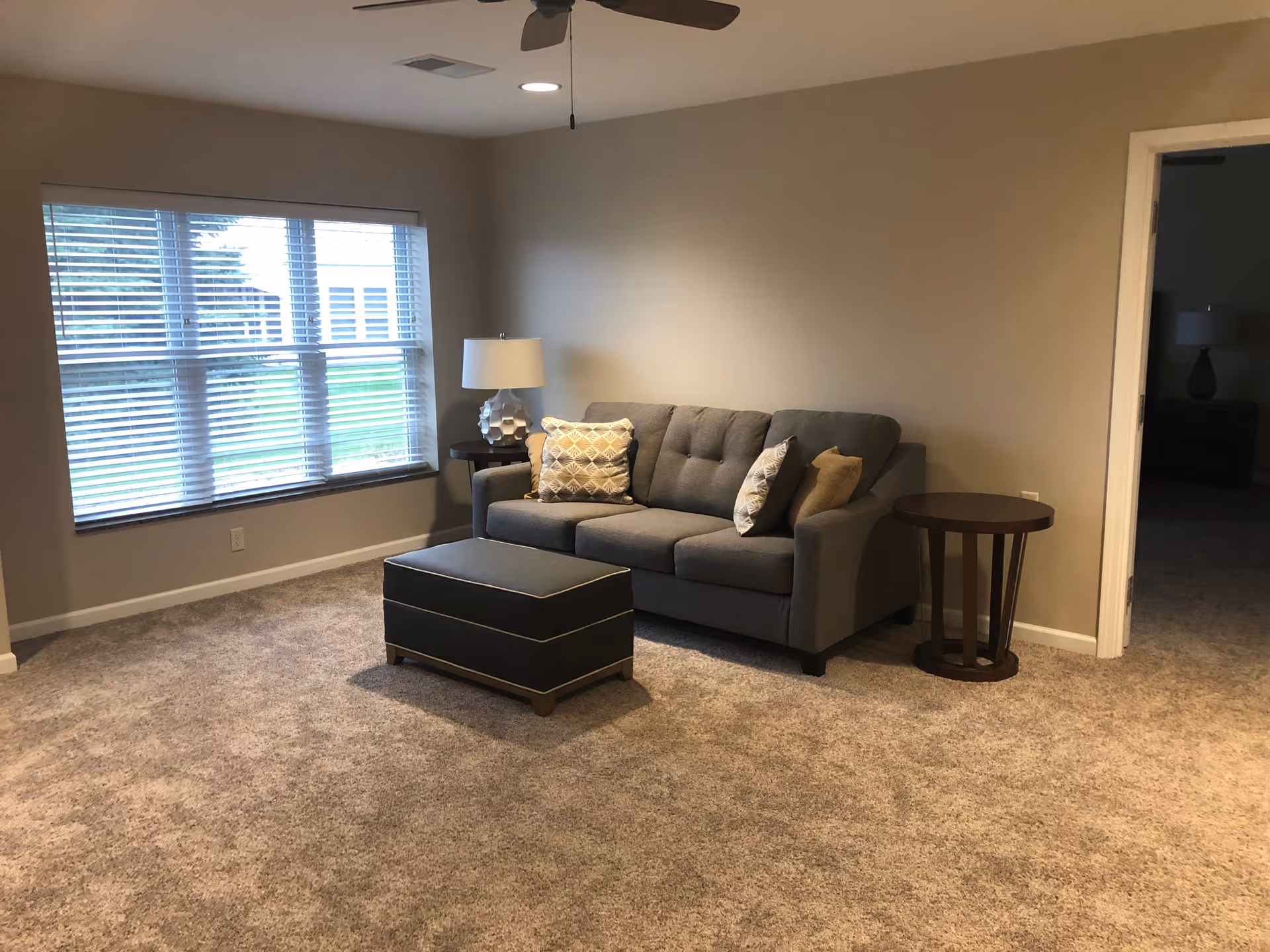 Carpeted living room with a gray sofa, ottoman, side tables and a large window with blinds.