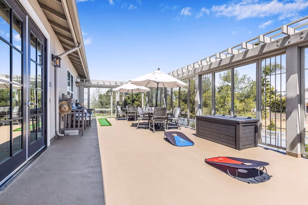 Spacious sunlit outdoor patio with tables, chairs and umbrellas under a pergola and two cornhole boards on the floor.