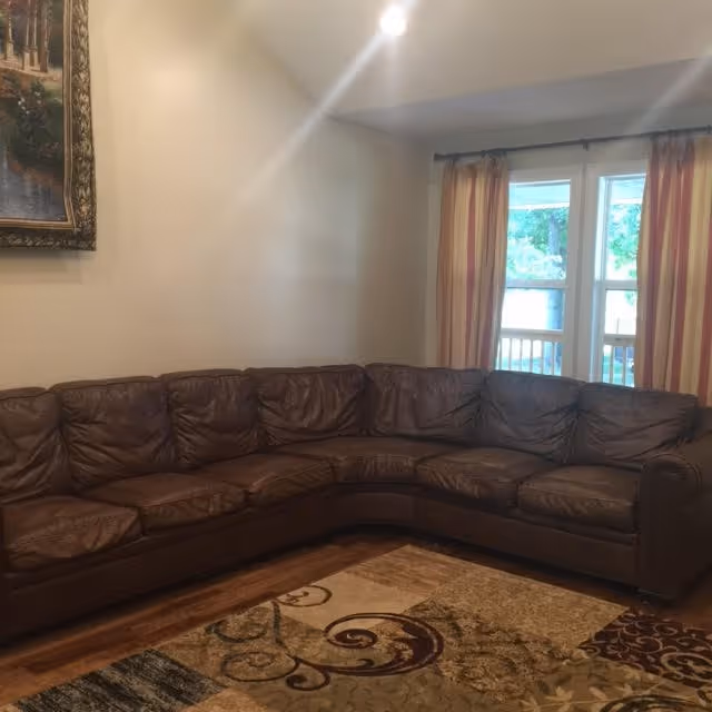A cozy living room with a large brown leather sectional sofa arranged in an L-shape. There is a patterned area rug on the wooden floor in front of the sofa. The room has beige walls, a window with striped curtains, and a framed painting hanging on the wall.