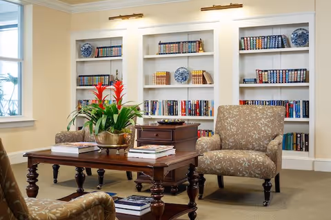 A cozy sitting area in a living room with two patterned armchairs and a wooden coffee table. The coffee table holds books and a decorative plant with red flowers. Behind the seating area are built-in white bookshelves filled with books and blue decorative plates. The room has beige walls and carpet, with a window on the left side letting in natural light.
