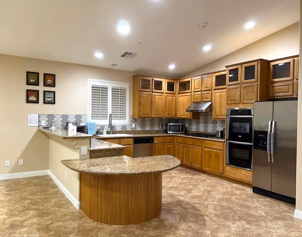 A spacious kitchen with wooden cabinets, granite countertops, and tiled flooring. The kitchen features a double oven, stainless steel refrigerator, microwave, dishwasher, and a sink under a window with white shutters. There are four framed pictures on the wall and recessed ceiling lights illuminating the area.