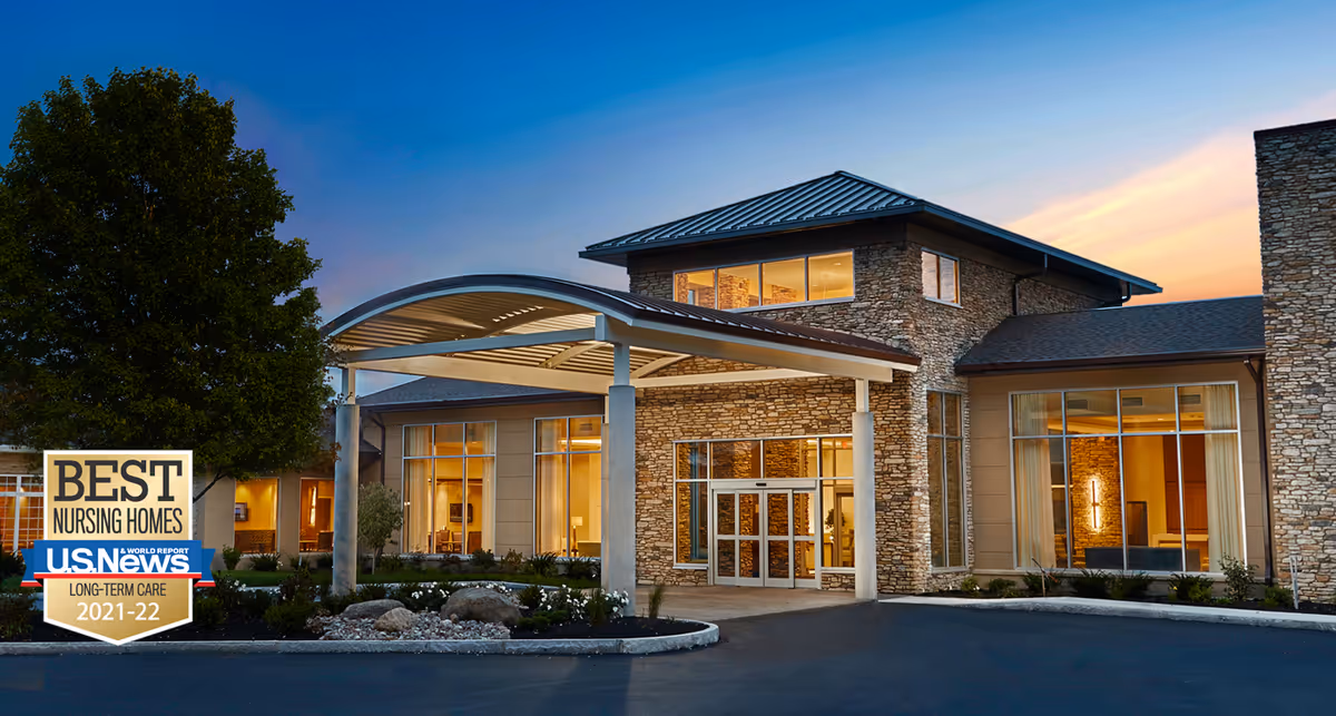Stone-clad nursing home front entrance at dusk with a covered porte-cochère and landscaping, with an award badge in the lower-left corner.