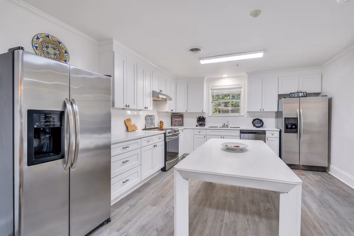 A modern kitchen with white cabinetry, two stainless steel refrigerators, a stove, dishwasher, and a white island table in the center. There is a window above the sink letting in natural light, and decorative items on the countertops and above the refrigerator.