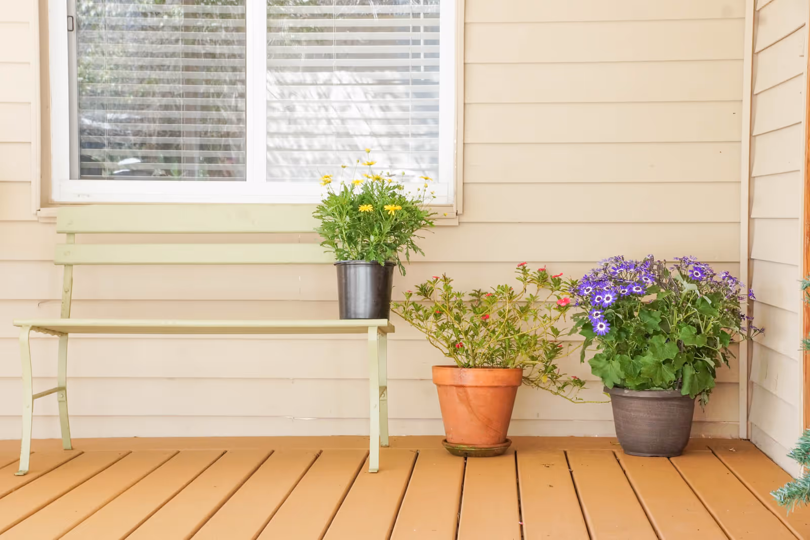 A beige front porch with a pale bench and several potted flowers on a wooden deck in front of a window.