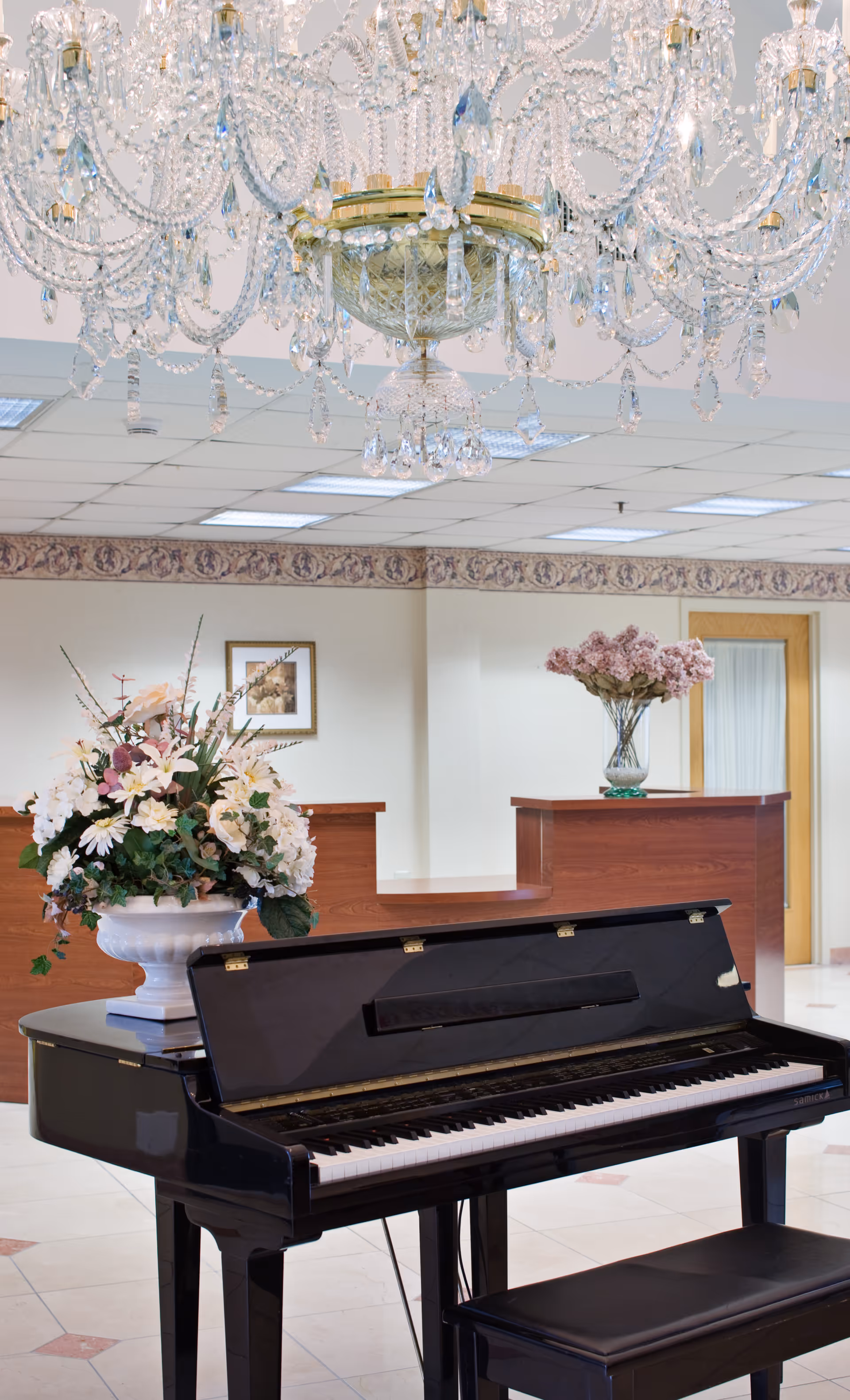 A grand piano is positioned in a decorated lobby under a crystal chandelier with floral arrangements and a reception desk in the background.