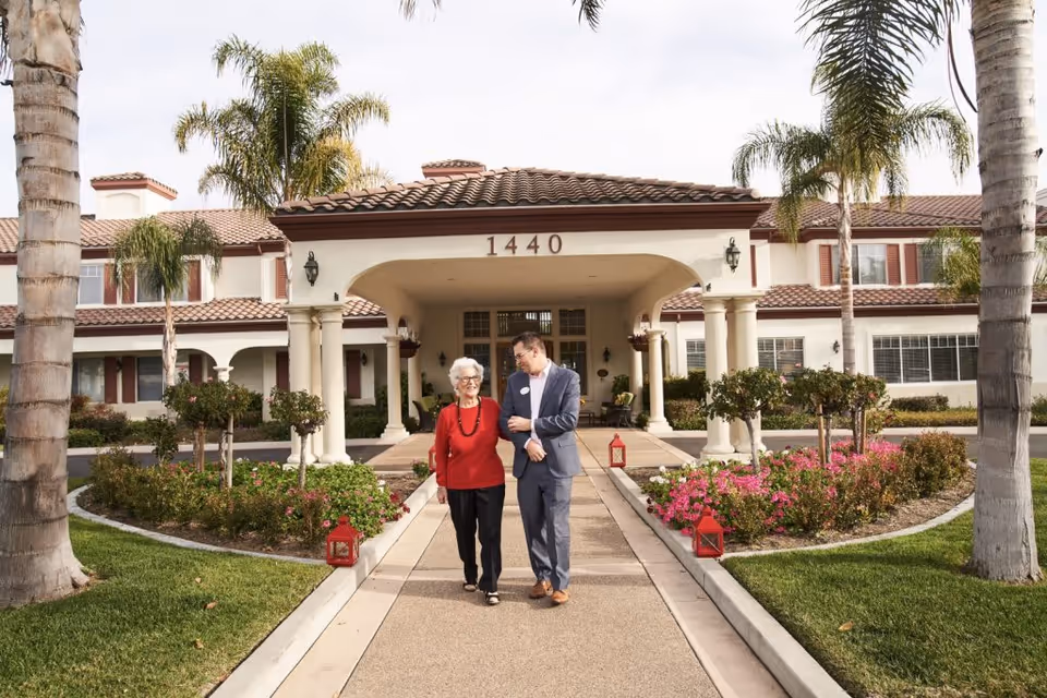 An elderly woman in a red sweater walking arm-in-arm with a man in a suit along a pathway leading to the entrance of a building with the number 1440 above the doorway. The building has a tiled roof, white columns, and is surrounded by palm trees and landscaped gardens with flowers.
