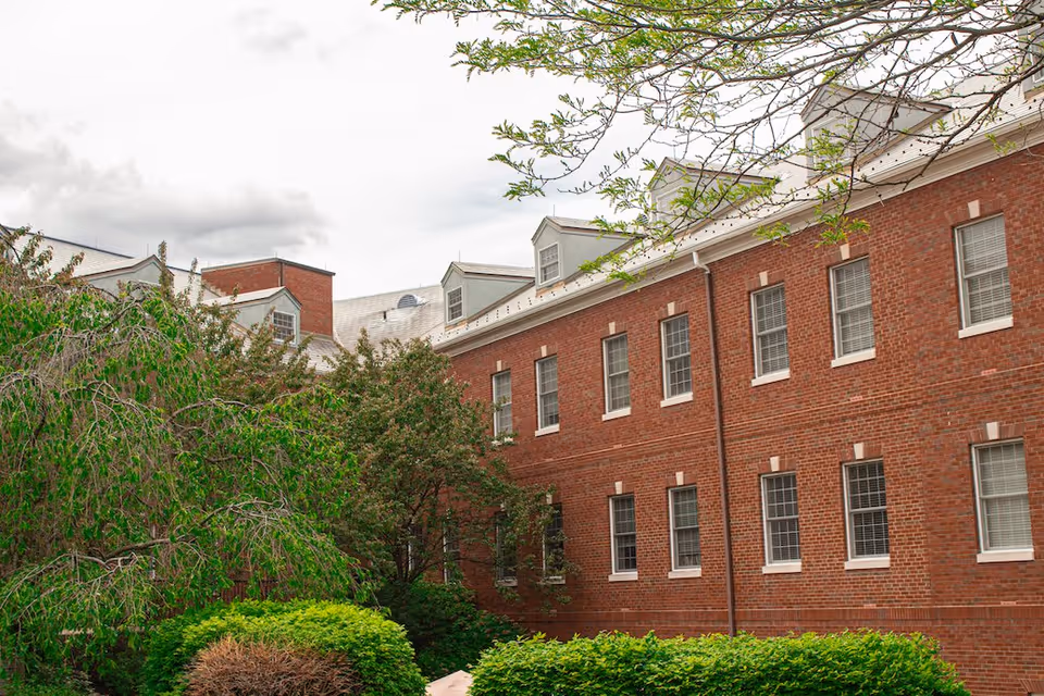 Exterior view of a two-story red brick building with multiple windows and dormer windows on the roof, surrounded by green trees and bushes under a cloudy sky.