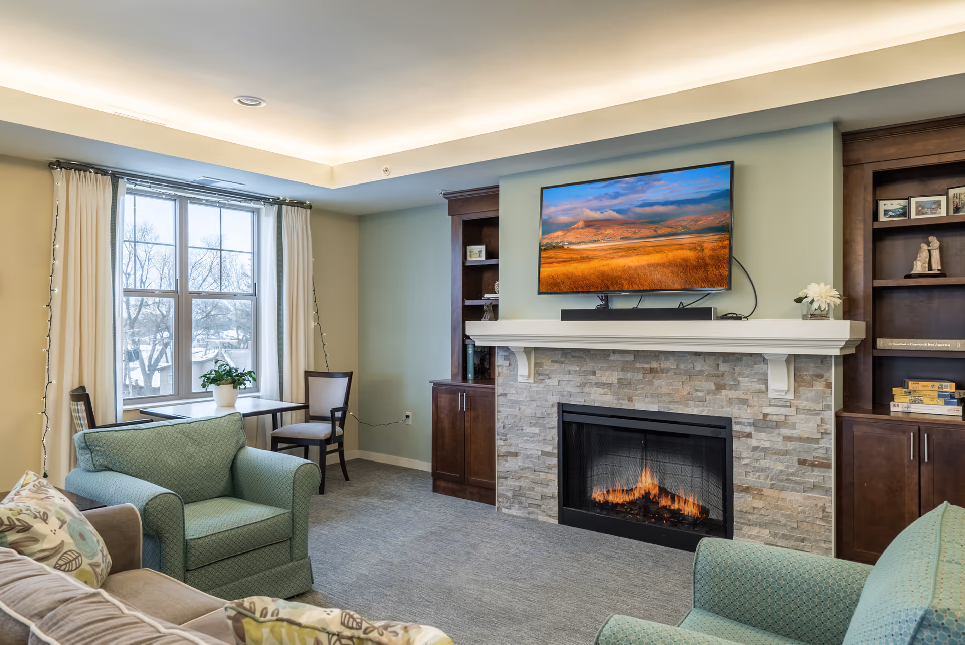 A cozy living room with a stone fireplace and a flat-screen TV mounted above it. The room has two green armchairs, a beige sofa with patterned cushions, a small table with two chairs near a window with cream curtains, and built-in dark wood shelves on either side of the fireplace.