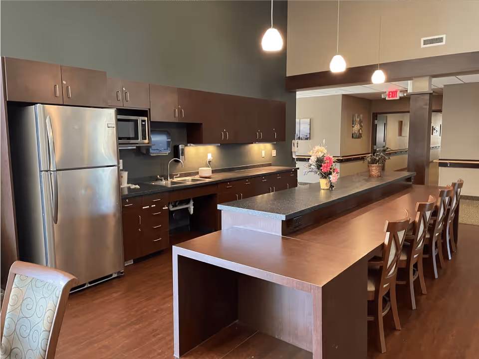 A modern kitchen area in a senior living facility featuring dark wood cabinets, a stainless steel refrigerator, a microwave, a sink, and a long counter with several wooden chairs. The space is well-lit with hanging pendant lights and decorated with small flower arrangements on the counter.