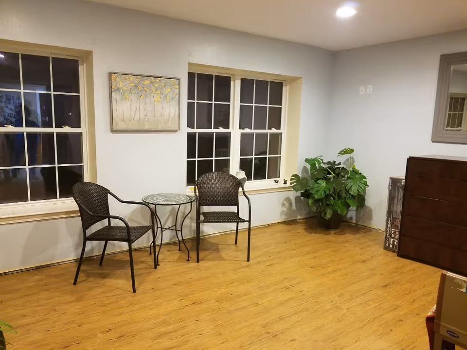 A small seating area with two black wicker chairs and a round glass-top table between them, placed in front of two large windows. A framed artwork with yellow leaves hangs on the wall above the table. To the right, there is a large green potted plant and a dark wooden dresser with a mirror above it. The floor is wooden, and the walls are painted light gray.