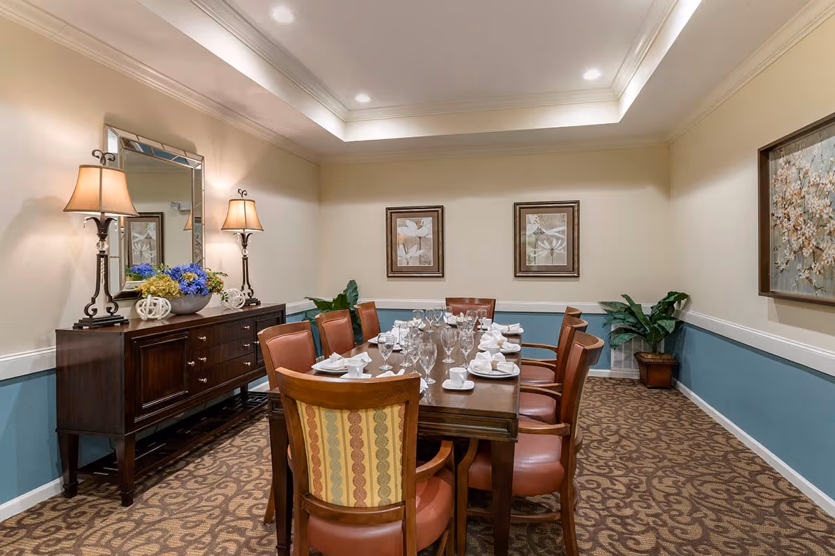 A formal dining room with a long wooden table set with white napkins, cups, and glassware. The room features eight chairs with leather seats and a patterned carpet. On the left side, there is a dark wooden sideboard with two table lamps and a decorative mirror above it. The walls are painted beige with a blue wainscoting and decorated with framed artwork and a potted plant in the corner.