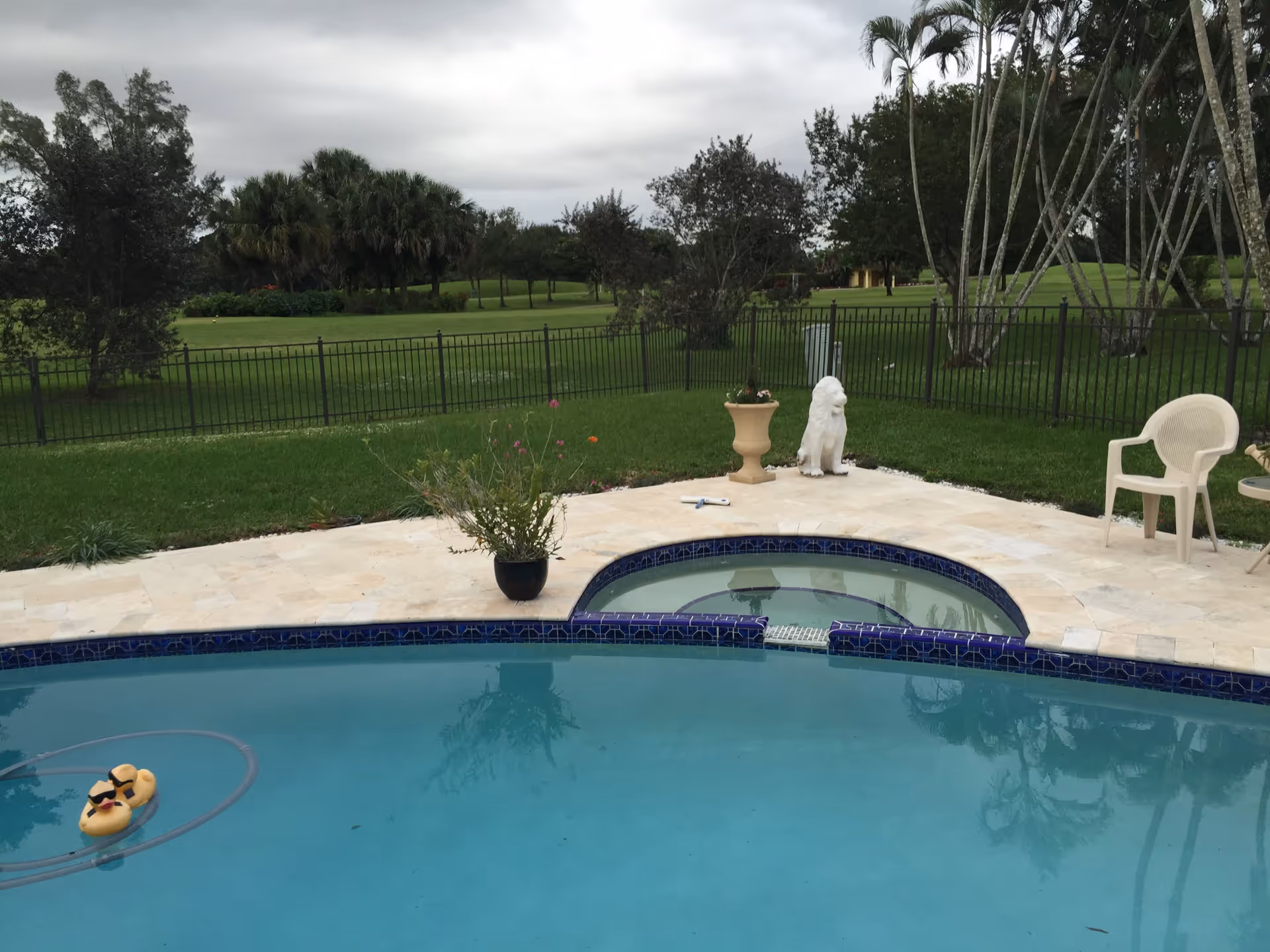 Outdoor view of a swimming pool with a small attached hot tub surrounded by a tiled deck. There is a potted plant on the deck, a white lion statue, a white plastic chair, and a small table. Beyond the pool area is a grassy lawn with trees and a black metal fence. The sky is cloudy.