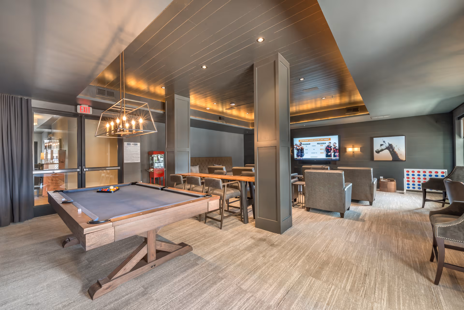 A modern recreational room featuring a pool table with balls arranged for a game, a long wooden table with chairs, a popcorn machine, a large flat-screen TV mounted on the wall, comfortable seating including armchairs and a bench, and a large Connect Four game near the window. The room has a warm lighting with a stylish ceiling and carpeted floor.