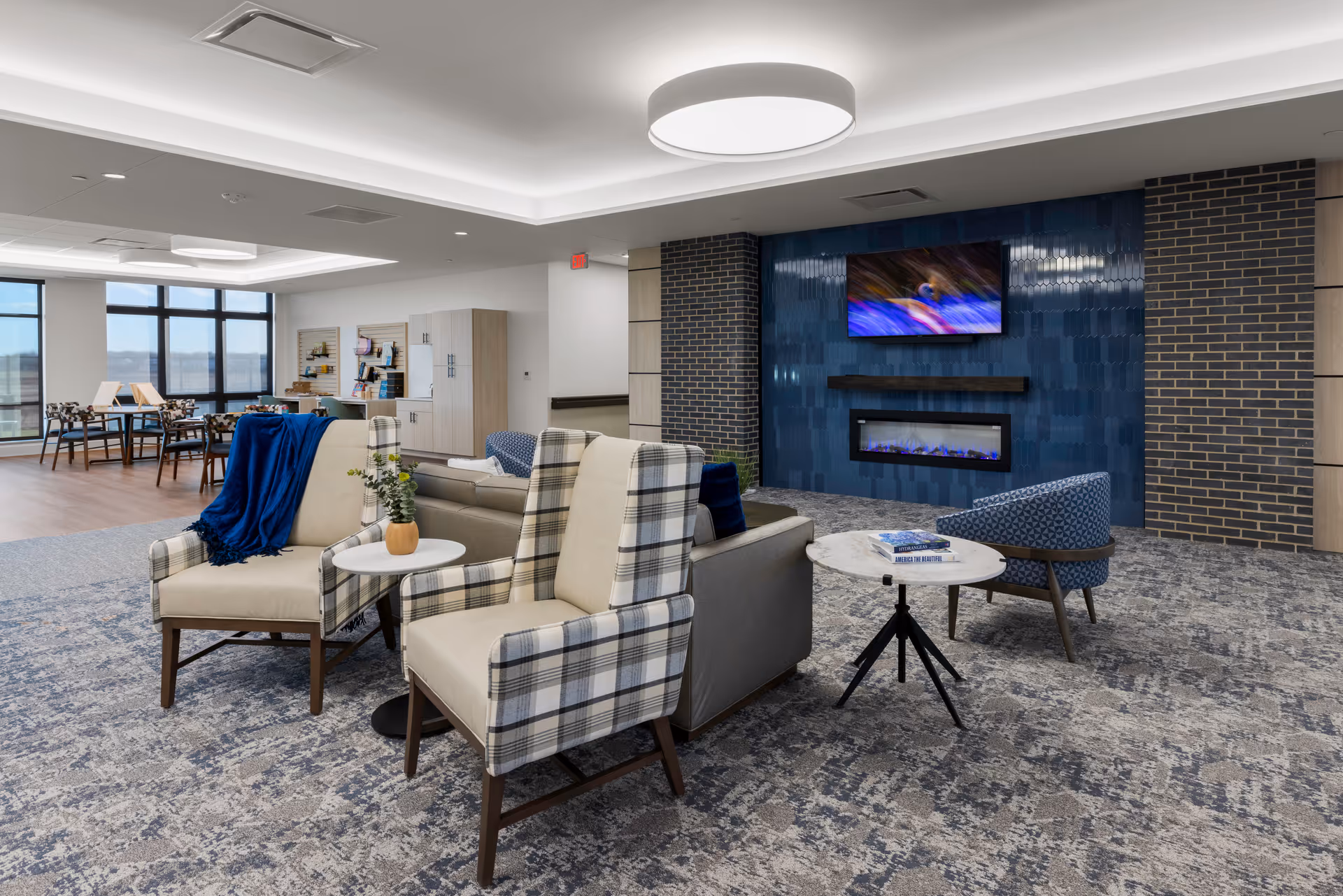 Modern communal lounge in a senior living facility featuring plaid armchairs, a sofa, small tables, a blue-tiled fireplace wall with a TV, and a dining area beyond large windows.