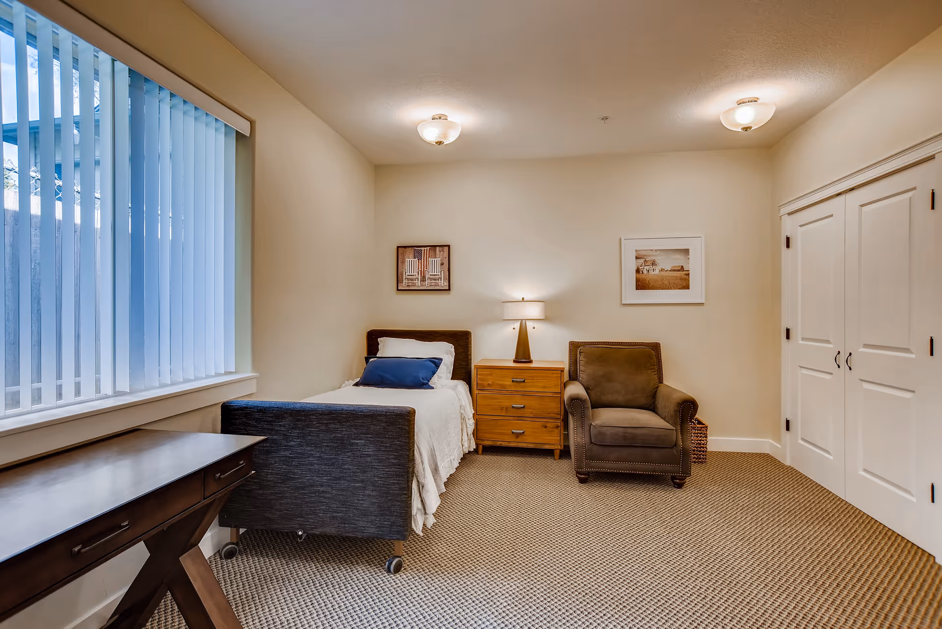 A cozy bedroom in a memory care facility featuring a single bed with white bedding and a blue pillow, a wooden nightstand with a lamp, a comfortable brown armchair, a wooden desk near a large window with vertical blinds, and two framed pictures on the beige walls.