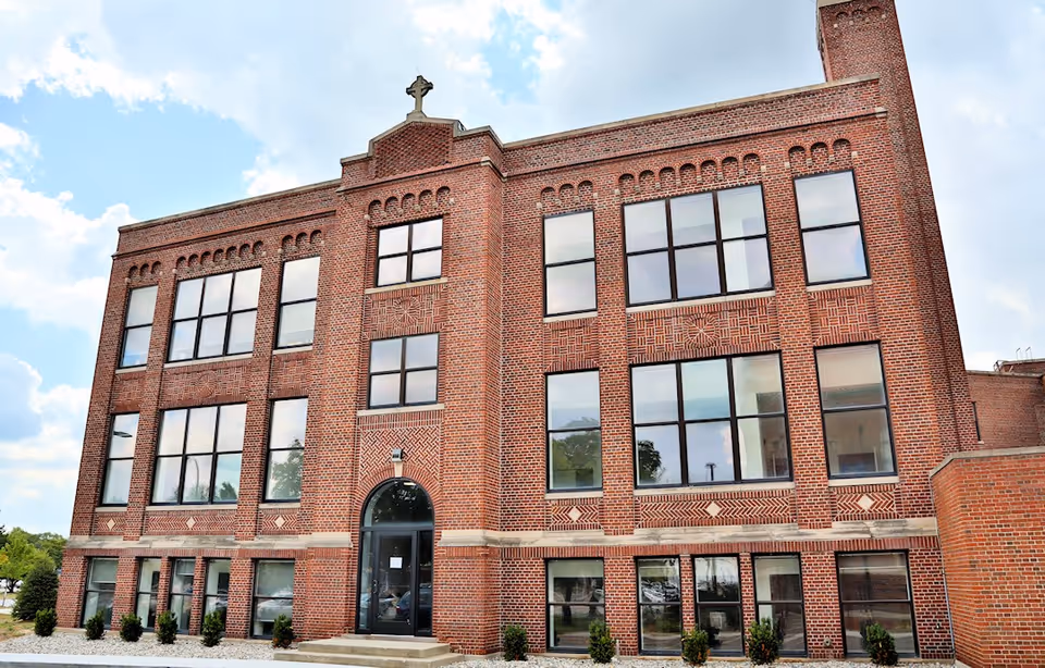 Front exterior of a three-story red brick building with large windows and a cross at the roof peak.