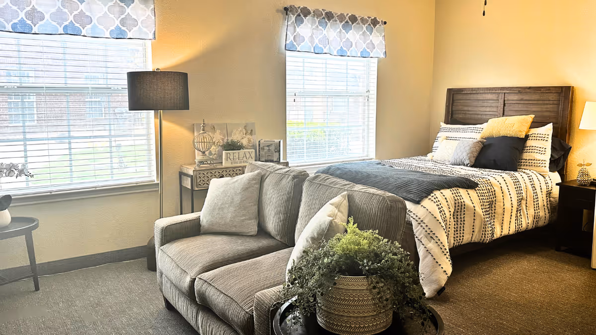 Sunlit bedroom with a bed, loveseat, floor lamp, side tables, and a potted plant by two large windows.