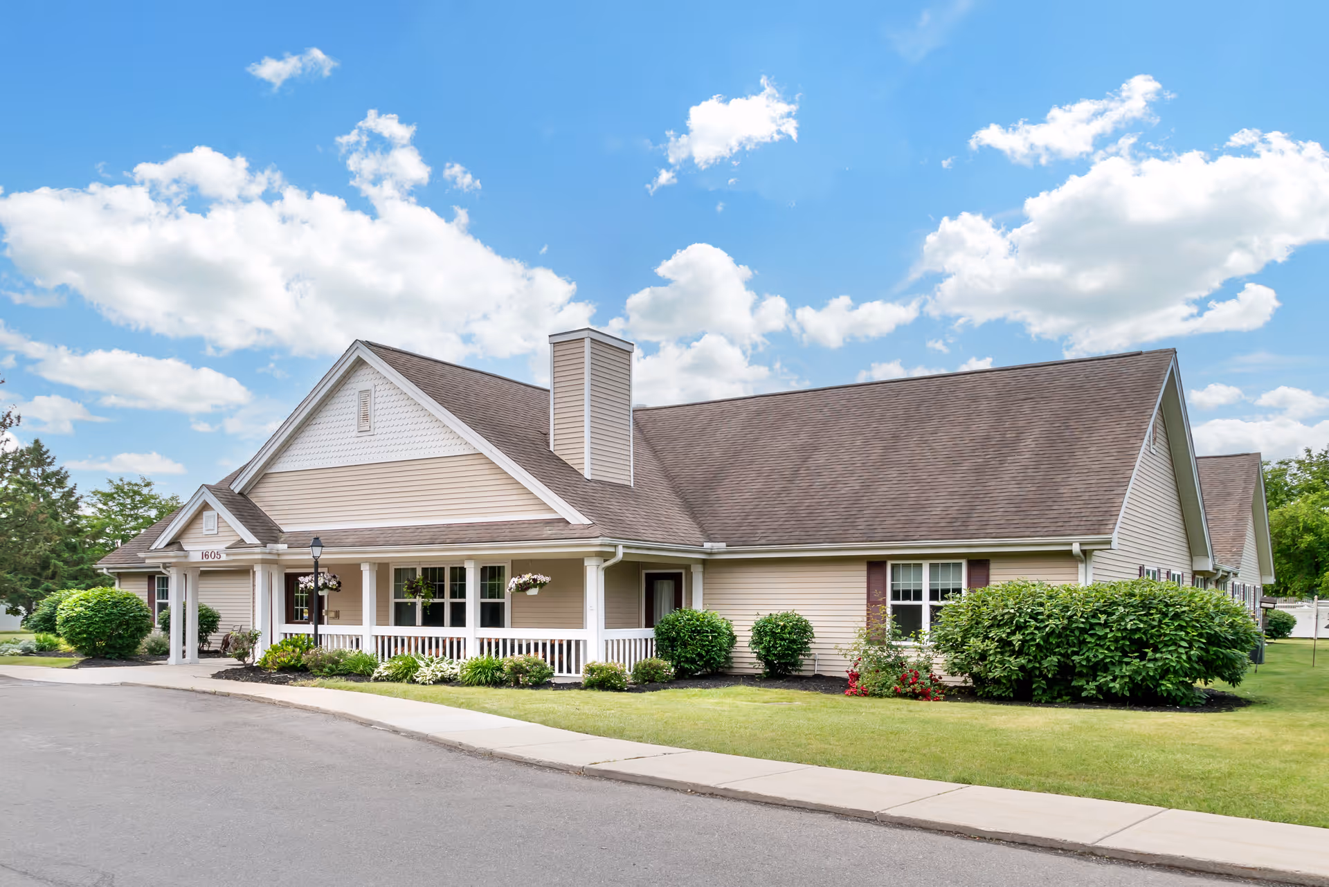 Exterior view of a single-story beige building with a brown shingled roof, white trim, and a covered porch with white railings. The building is surrounded by green grass, bushes, and flowers under a partly cloudy blue sky.