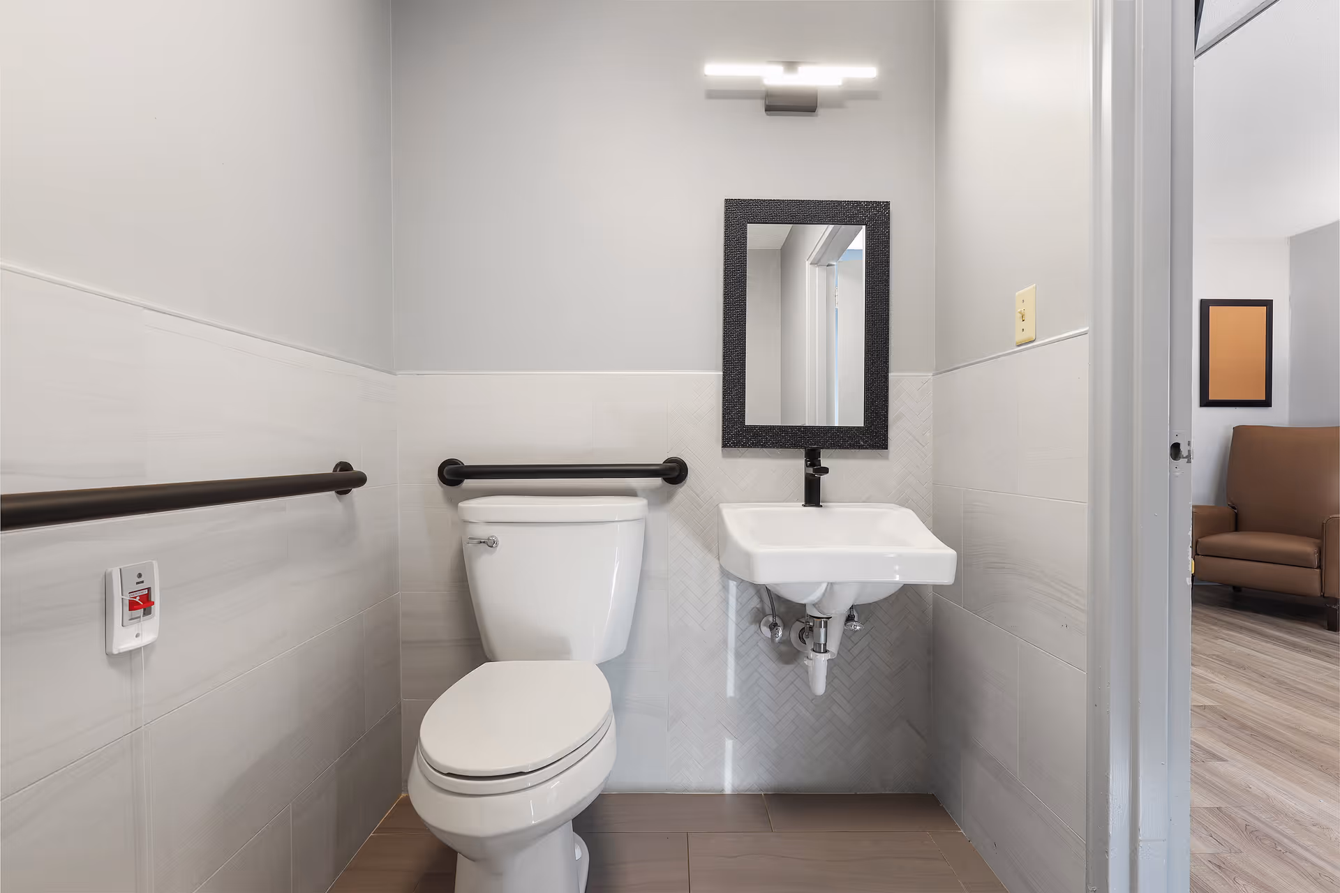 Accessible bathroom with a toilet, wall-mounted sink and mirror, horizontal grab bars, and an emergency call button.