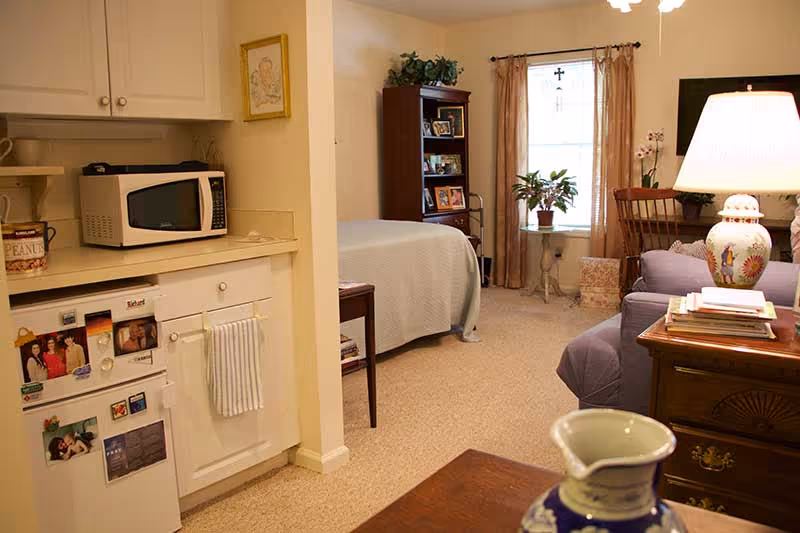 Studio-style interior showing a small kitchenette open to a living/bedroom area with a bed, bookshelf, sofa, and lamp.