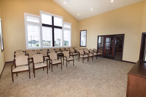 A spacious room with beige walls and carpeted floor featuring two rows of beige cushioned chairs with wooden armrests arranged facing a large window. The window has multiple panes and offers a view of houses outside. There are two dark-framed glass doors on the right wall and a wooden cabinet or table on the right side of the image.