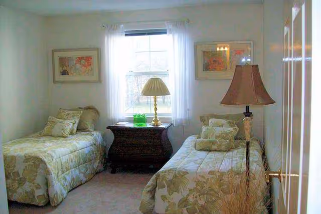 A bedroom with two twin beds on either side of a window. Each bed has floral patterned bedding and multiple pillows. Between the beds is a dark wooden nightstand with a brass lamp and a green glass decorative item. The window has sheer white curtains, and there are framed floral artworks on the walls above each bed. A floor lamp with a beige shade is positioned near the right bed.
