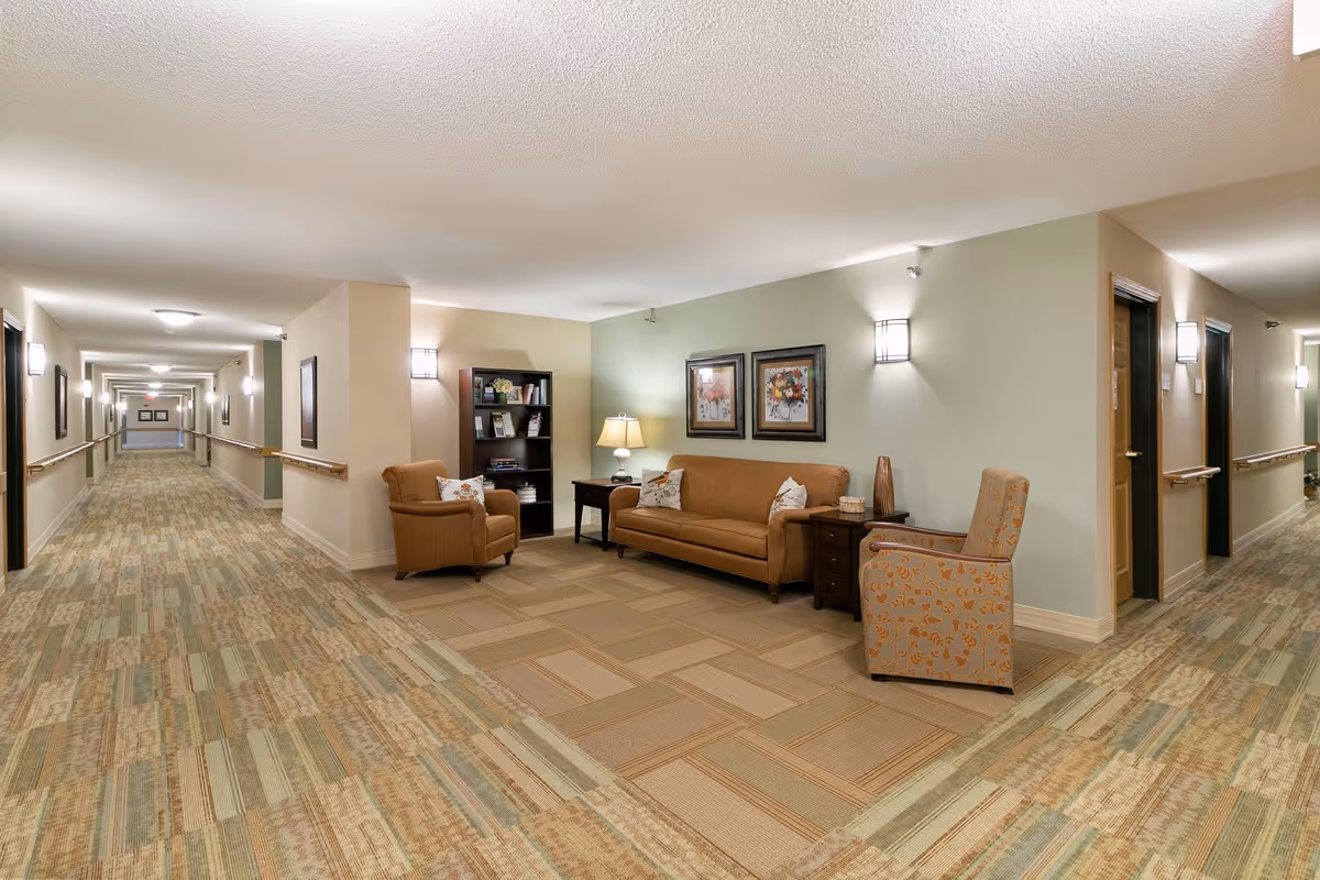 A well-lit hallway in a senior living facility with patterned carpet and handrails along the walls. There is a cozy seating area with a brown sofa, two armchairs, a side table with a lamp, a bookshelf, and two framed floral paintings on the wall.