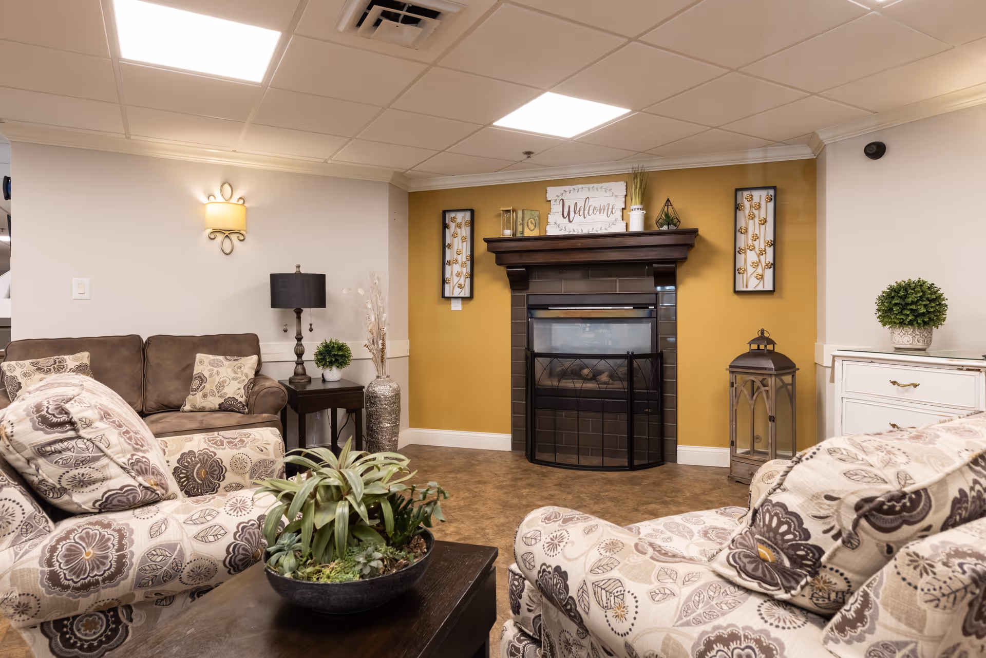 A cozy living room area with patterned armchairs and a brown sofa. There is a dark wooden coffee table with a plant centerpiece. The room features a fireplace with a dark wooden mantle, decorated with a 'Welcome' sign and small decorative items. The walls are painted white and mustard yellow, with framed floral artwork on either side of the fireplace. Additional decor includes a table lamp, a large vase with dried plants, a lantern, and a small potted plant on a white cabinet.