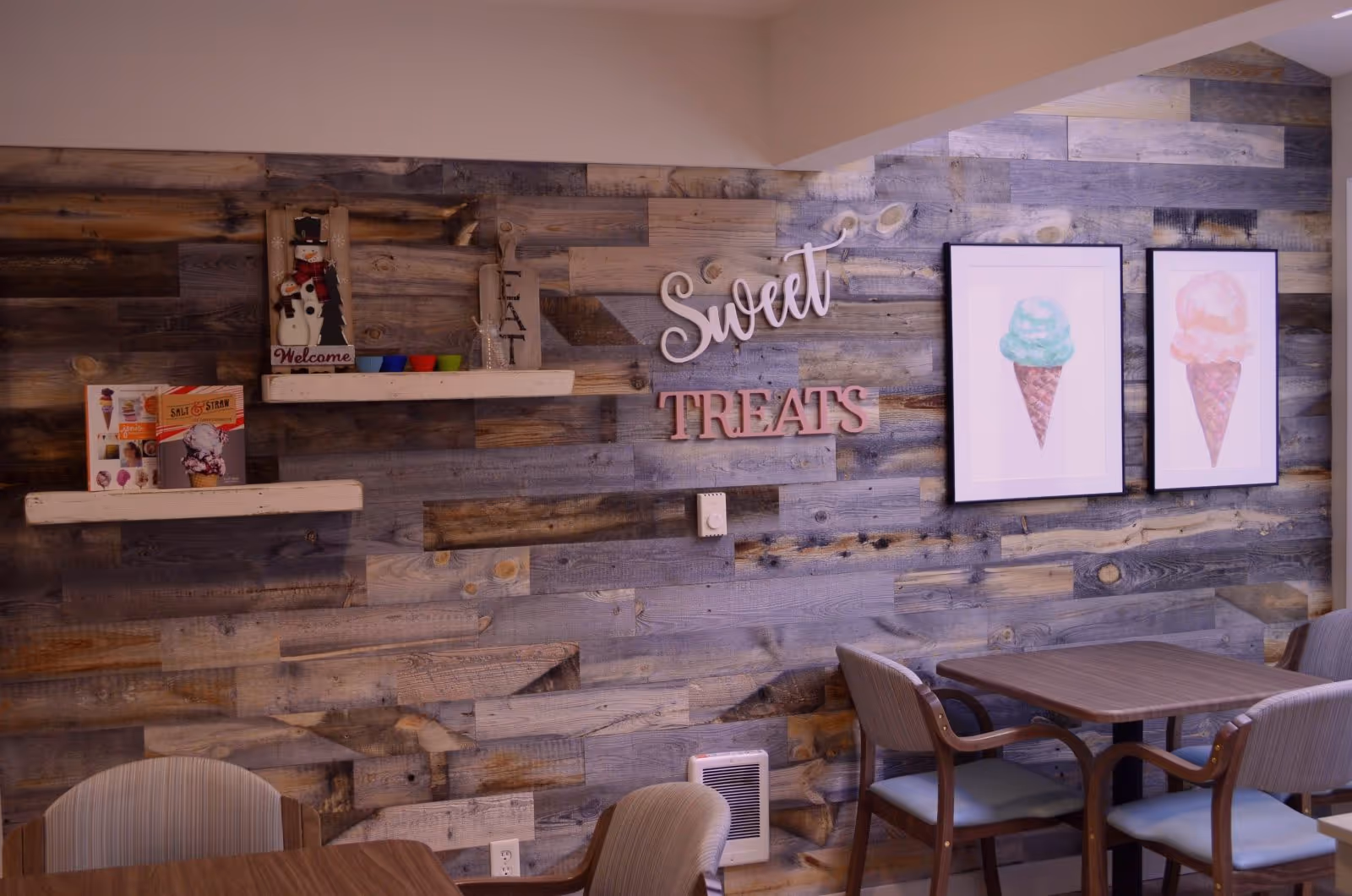 A cozy dining area with a rustic wooden plank wall decorated with two framed pictures of ice cream cones, a wooden sign that says 'Sweet TREATS', and two white shelves holding colorful cups, a snowman decoration, and menus. The room has a wooden table with four cushioned chairs.