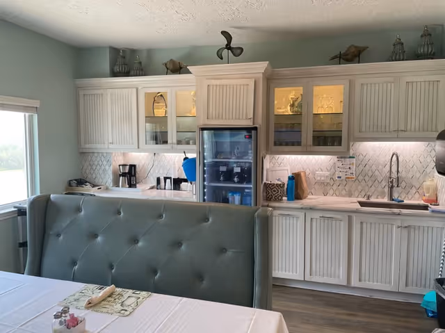 Interior view of a kitchen area with white cabinets, a glass-front refrigerator, a coffee maker, and a sink. There is a cushioned bench with button tufting in the foreground and a table with a white tablecloth and placemats. Decorative items are placed on top of the cabinets.