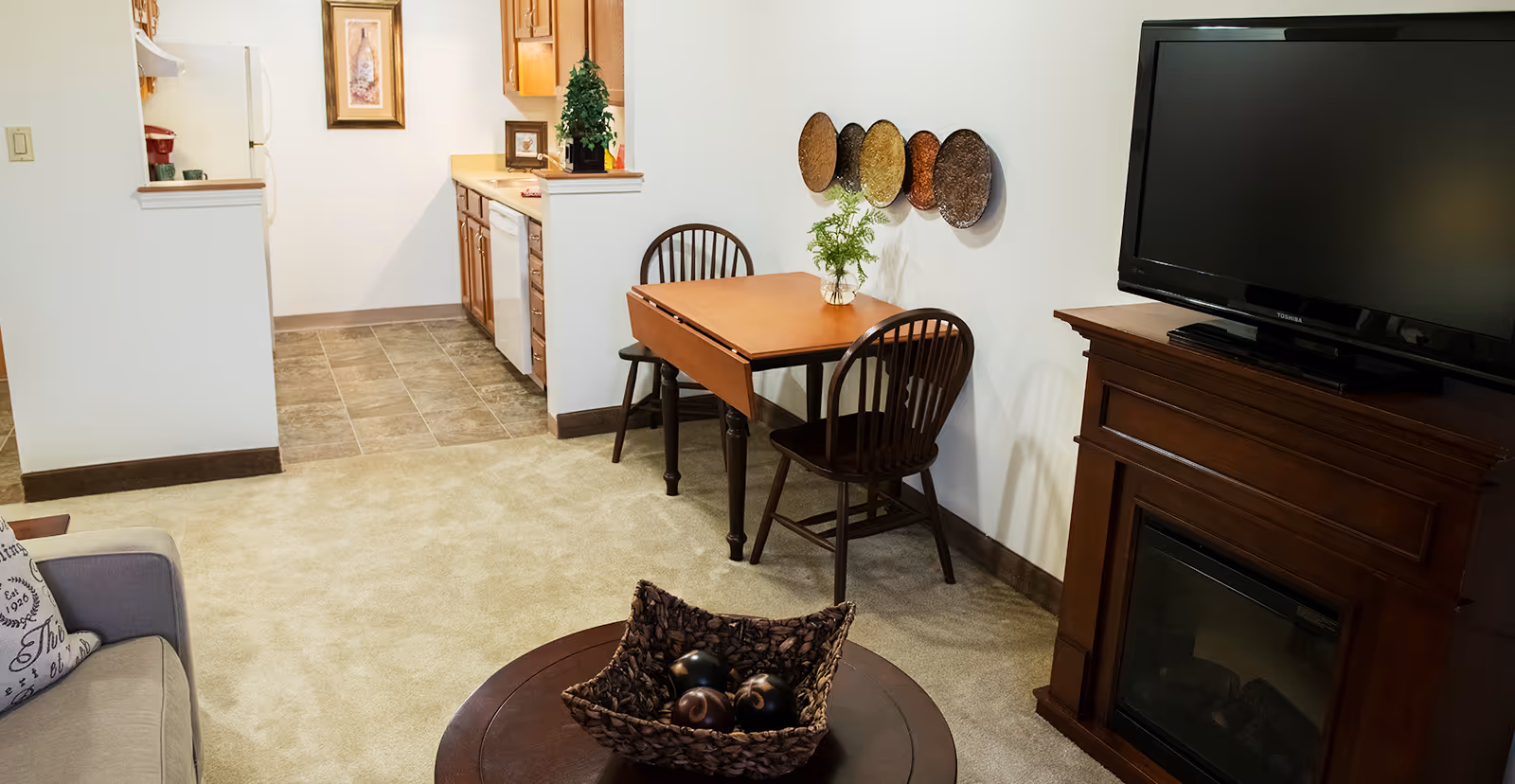 A cozy living area with a small wooden dining table and two chairs against a white wall decorated with four round woven wall hangings. To the right, there is a dark wooden TV stand with a flat-screen TV on top. In the foreground, a round coffee table holds a woven basket with decorative balls. The living area carpet transitions to a tiled kitchen floor with wooden cabinets, a white refrigerator, and a dishwasher visible.