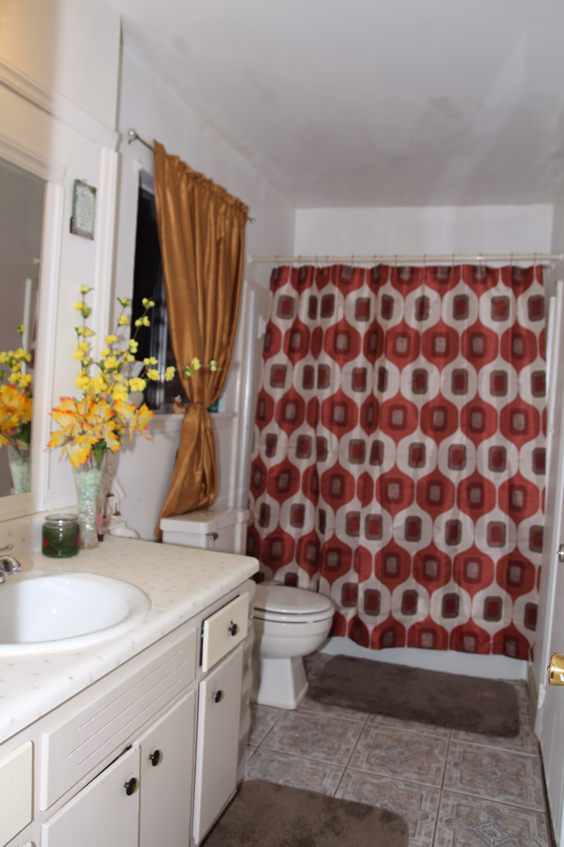 A bathroom with a white sink and countertop, white cabinets below, a toilet, and a shower with a red and white patterned curtain. There is a brown curtain on the window and a vase with yellow and orange flowers on the countertop. The floor is tiled and there are brown bath mats on the floor.