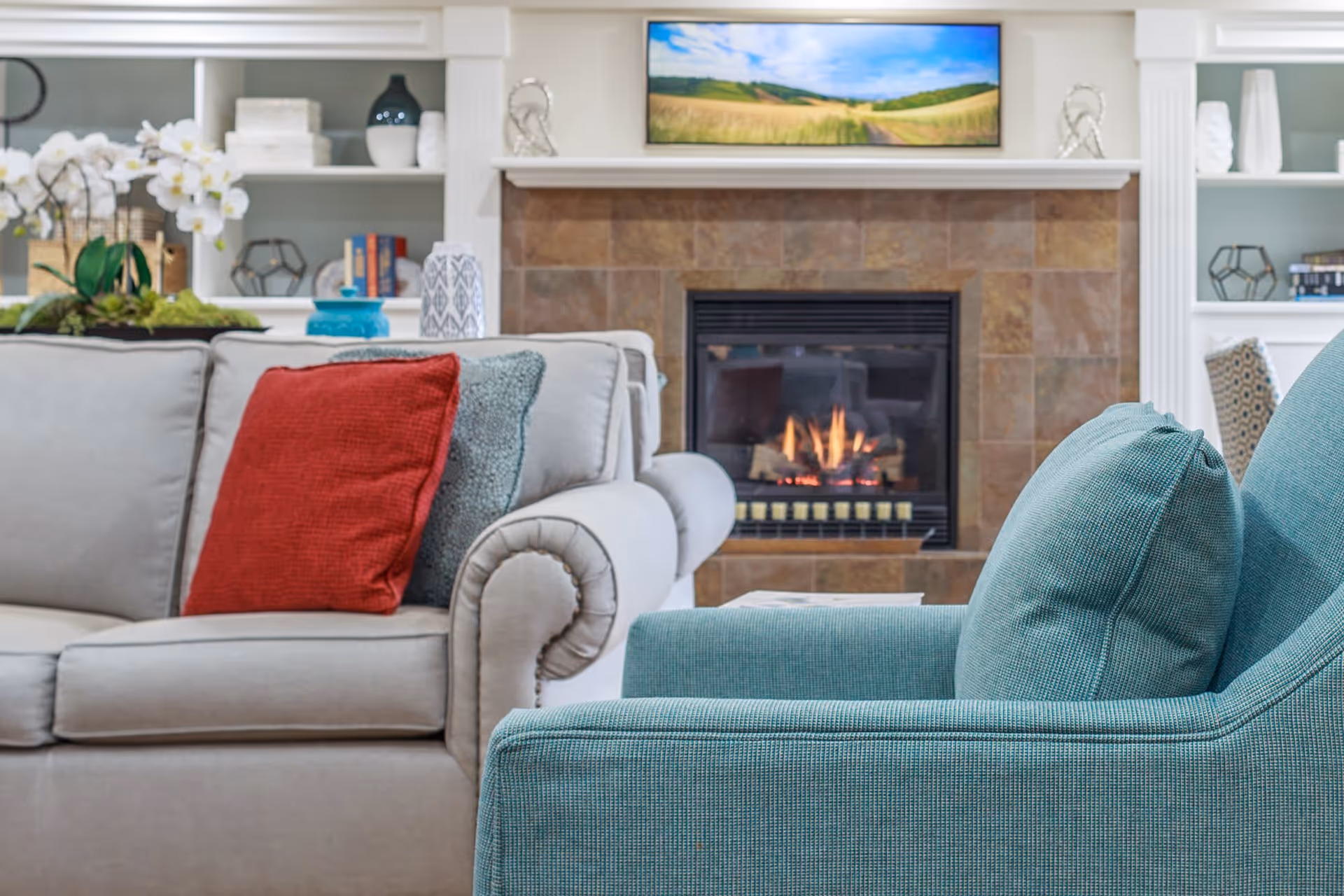 A cozy living room with a beige sofa adorned with red and gray cushions, a teal armchair, a lit fireplace with a tiled surround, and built-in white shelves decorated with books, vases, and plants.